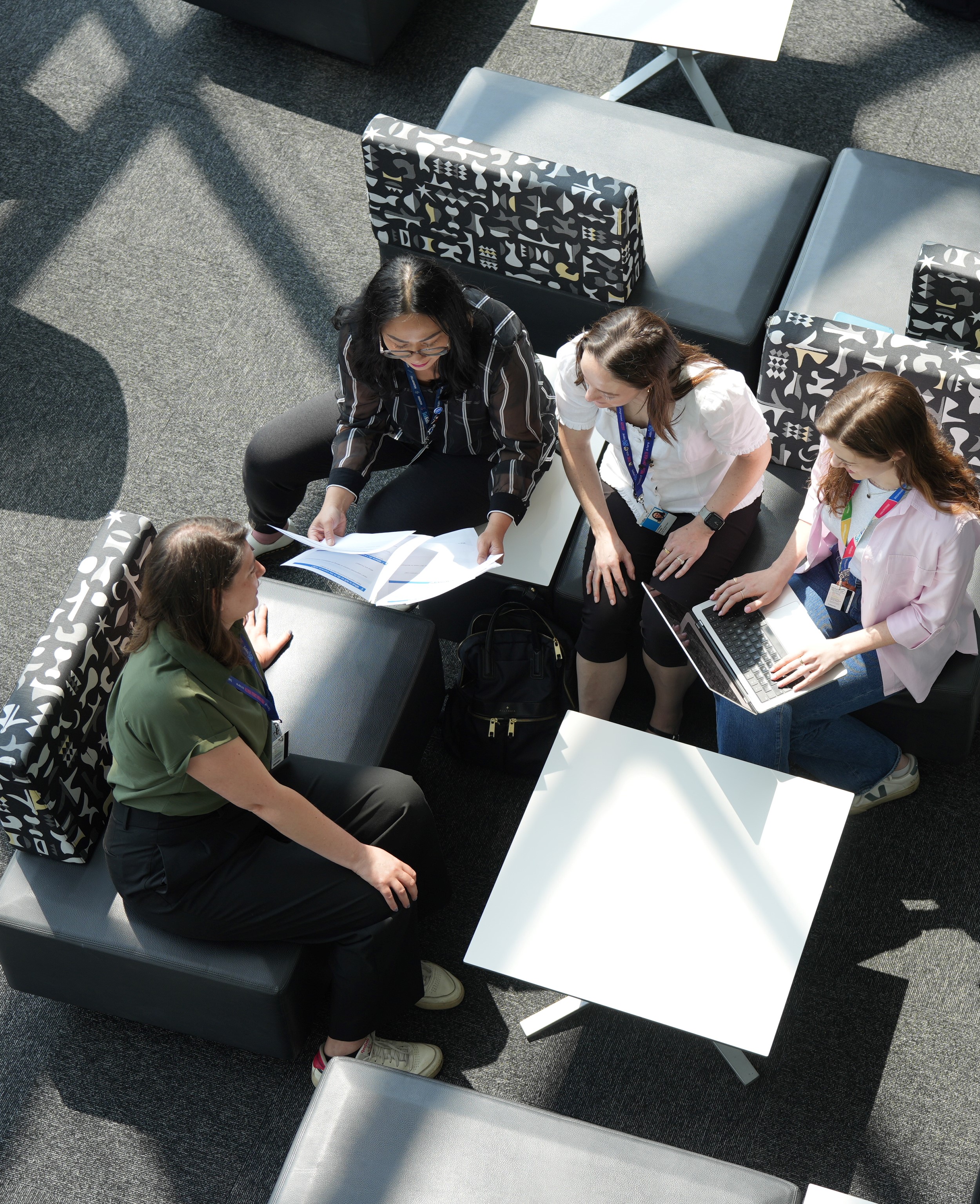 Aerial view of four women collaborating in a modern office space, sitting on black patterned chairs around a white coffee table with documents and a laptop, wearing conference lanyards in a sunlit room with geometric shadows.