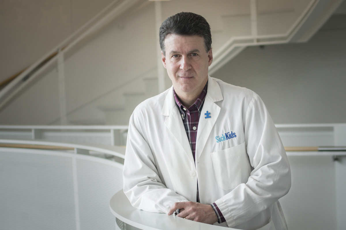 Dr. Stephen Scherer in a white SickKids lab coat standing with arms crossed in a laboratory, with researchers and scientific equipment visible in the background
